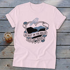 Pink t-shirt with a heart-shaped design featuring a bird and mountain landscape, text reads "HEART IN THE HOLLER MILES CAN'T MEASURE IT".