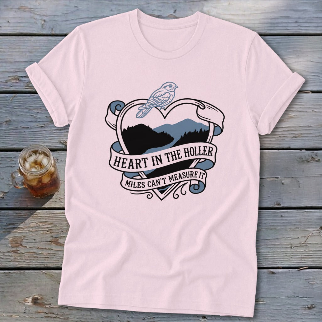Pink t-shirt with a heart-shaped design featuring a bird and mountain landscape, text reads "HEART IN THE HOLLER MILES CAN'T MEASURE IT".