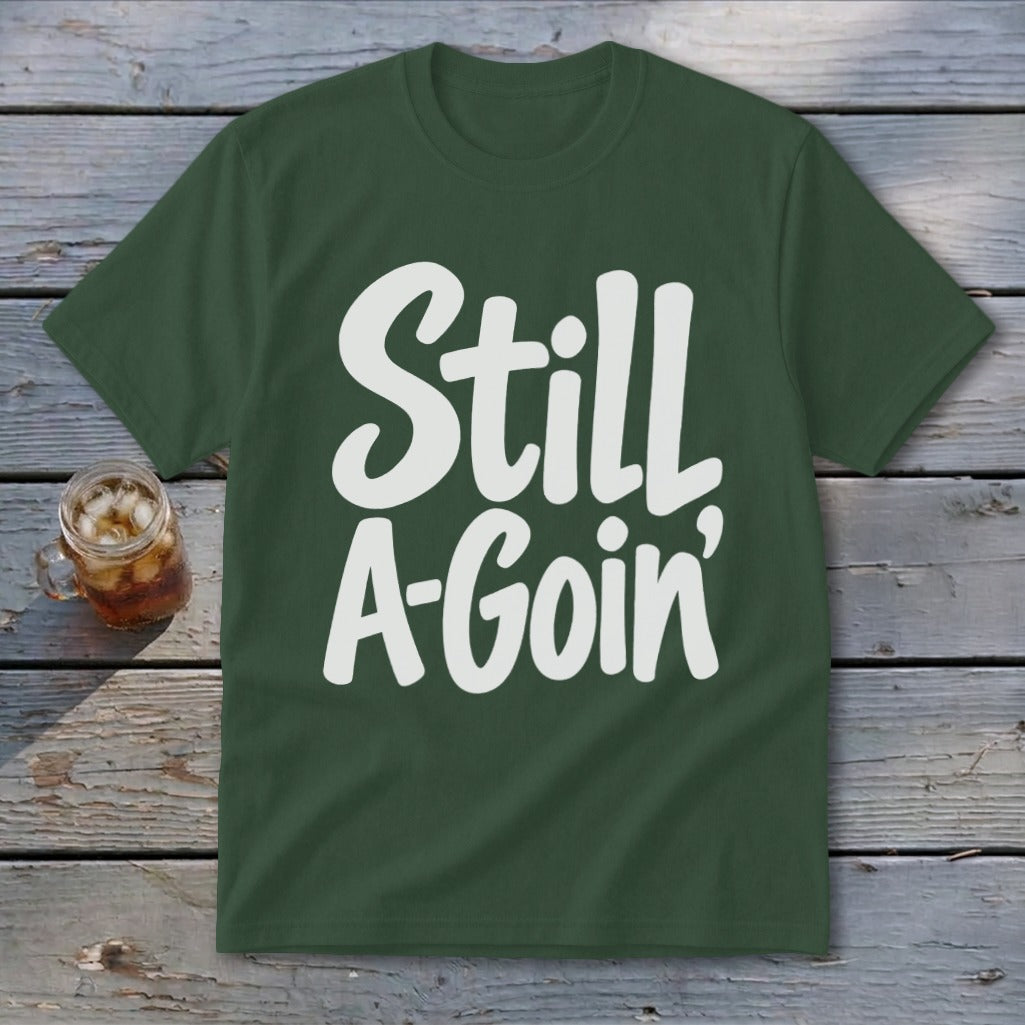 A dark green t-shirt featuring the white text Still A-Goin displayed on a weathered wooden surface.