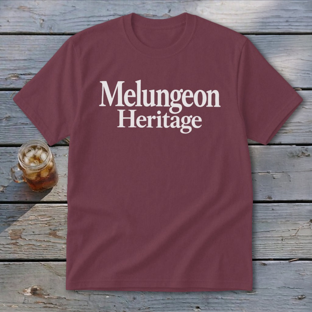 A maroon short-sleeved t-shirt with white text reading "Melungeon Heritage" centered on the chest, laid flat on a rustic wooden table next to a glass of iced tea.