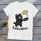 A white t-shirt featuring a cartoon black bear flipping a pancake with the text hoecakes.