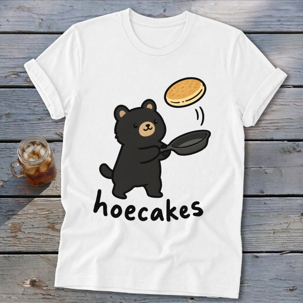 A white t-shirt featuring a cartoon black bear flipping a pancake with the text hoecakes.