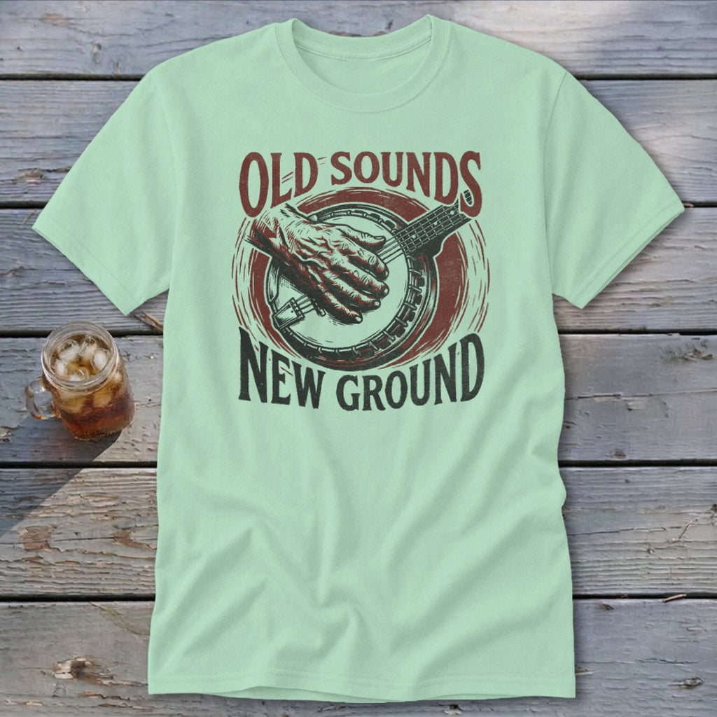 A light green t-shirt featuring a banjo graphic and the text "Old Sounds New Ground" on a wooden background.