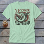 A light green t-shirt featuring a banjo graphic and the text "Old Sounds New Ground" on a wooden background.