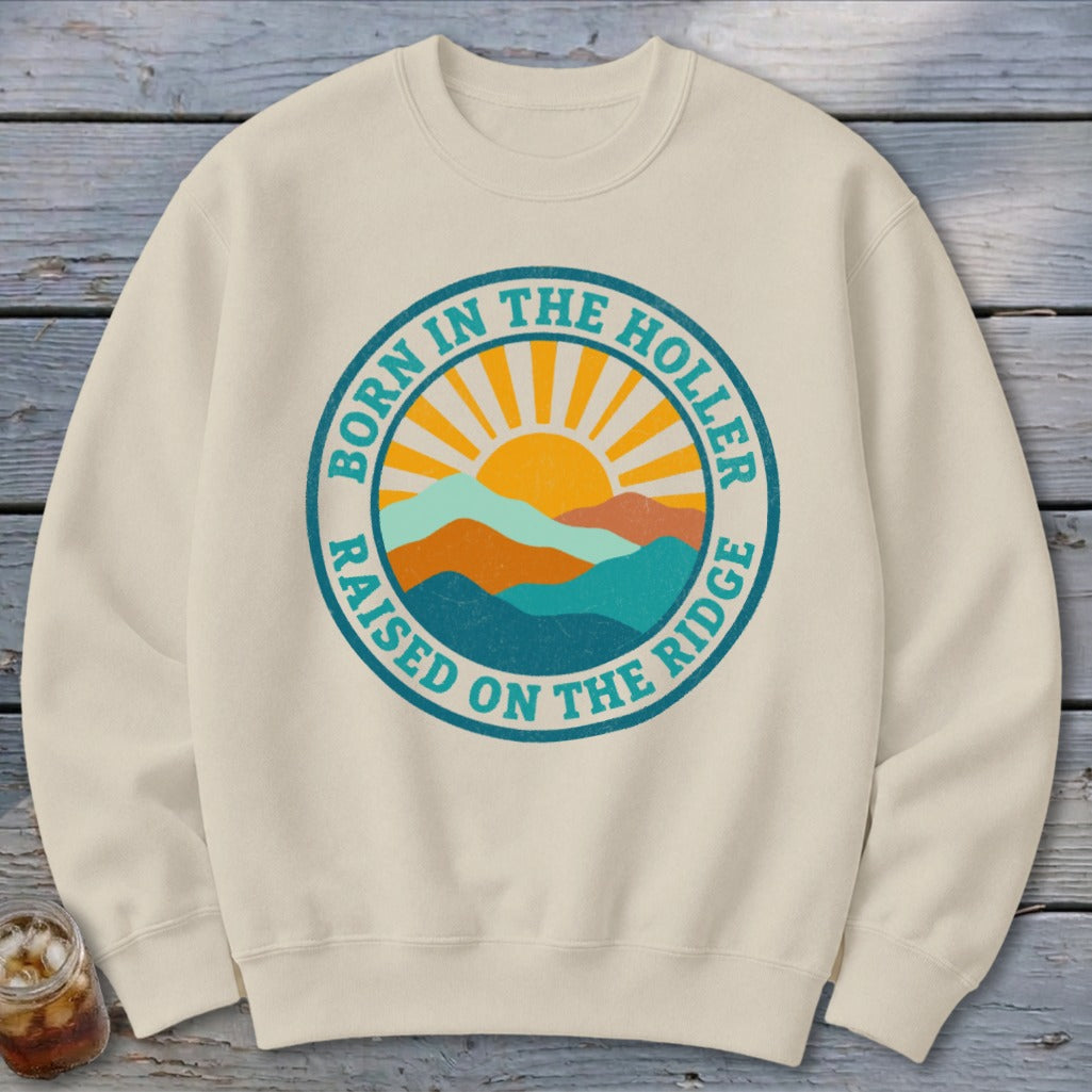 A cream crewneck sweatshirt featuring a circular graphic with mountains, a rising sun, and text that reads BORN IN THE HOLLER RAISED ON THE RIDGE.