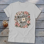 Light grey t-shirt with "Mountain Mama Raised Em Right" text within a floral wreath design.
