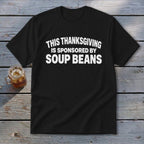 Black t-shirt featuring the white text "THIS THANKSGIVING IS SPONSORED BY SOUP BEANS" resting on a wooden surface.