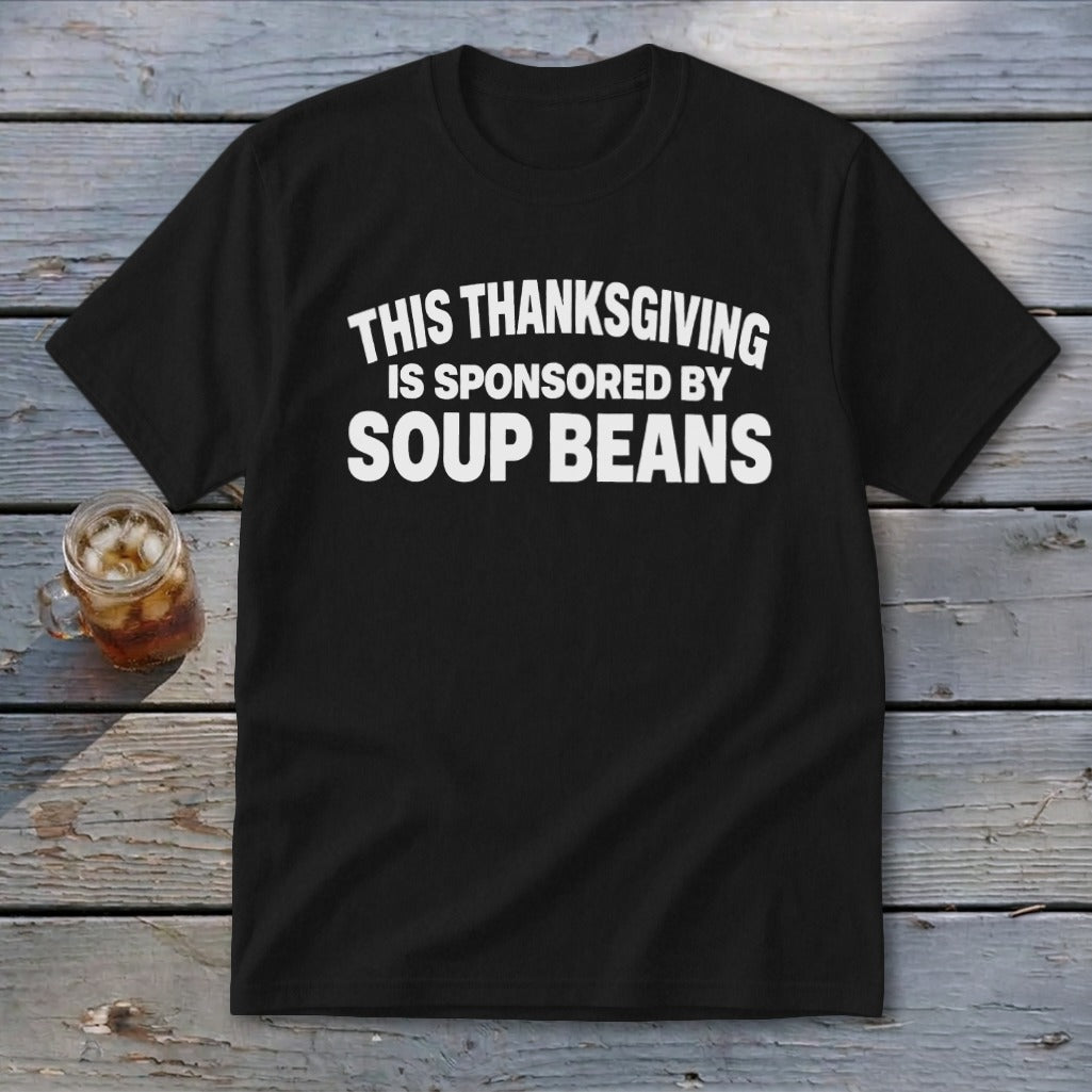 Black t-shirt featuring the white text "THIS THANKSGIVING IS SPONSORED BY SOUP BEANS" resting on a wooden surface.