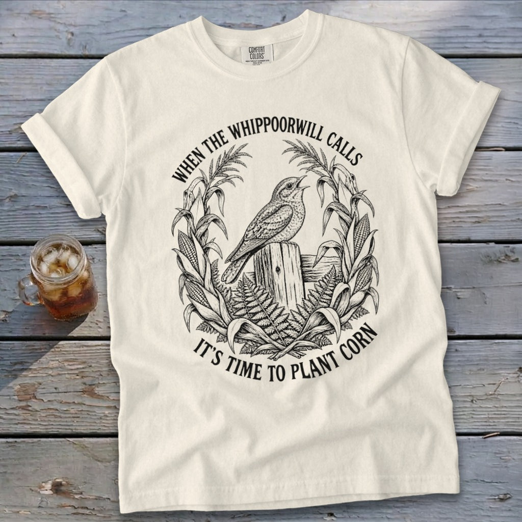 An off-white t-shirt featuring a black line art graphic of a whippoorwill perched on a wooden post, surrounded by corn stalks and ferns, with the text WHEN THE WHIPPOORWILL CALLS IT'S TIME TO PLANT CORN.