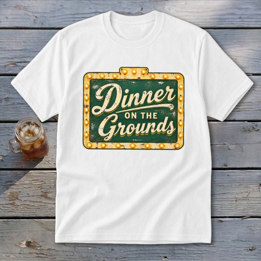A white t-shirt featuring a green vintage marquee sign graphic that reads Dinner on the Grounds.