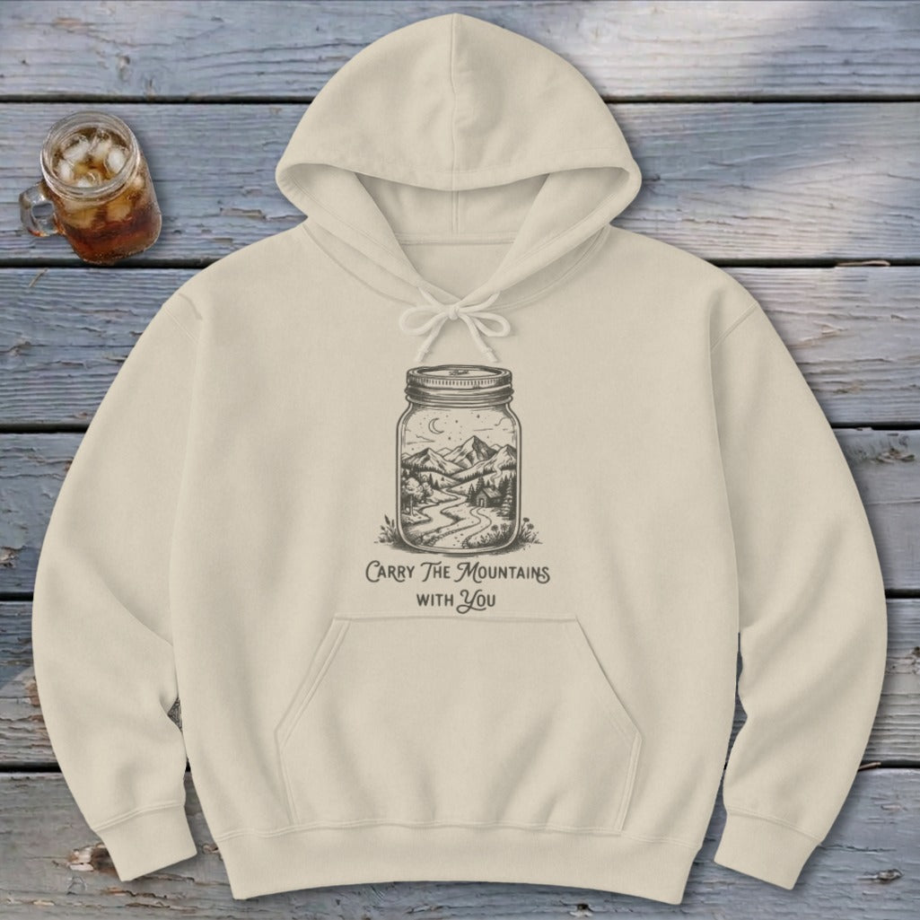 A cream-colored hoodie features a graphic of mountains in a jar with the text Carry The Mountains With You.