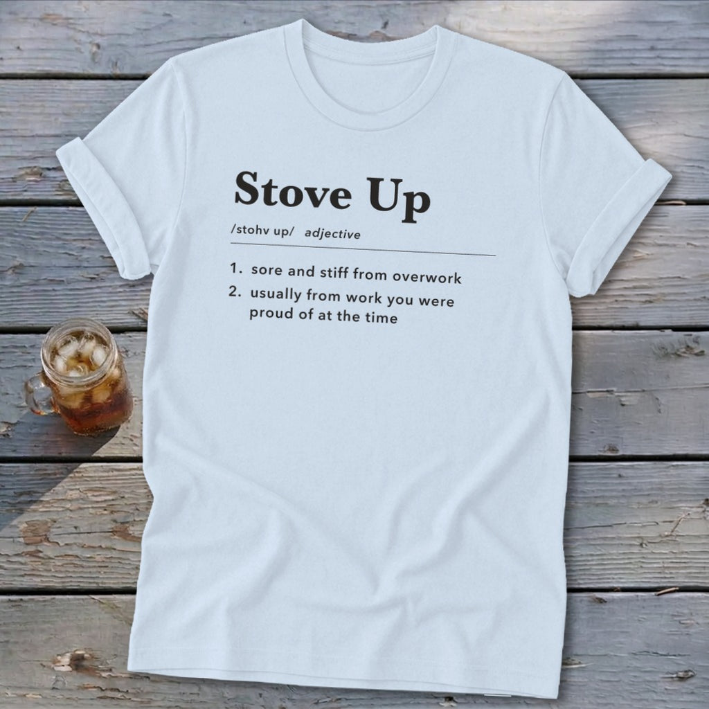 A light blue crewneck t-shirt rests on a rustic wooden table, featuring a humorous dictionary definition of Stove Up.