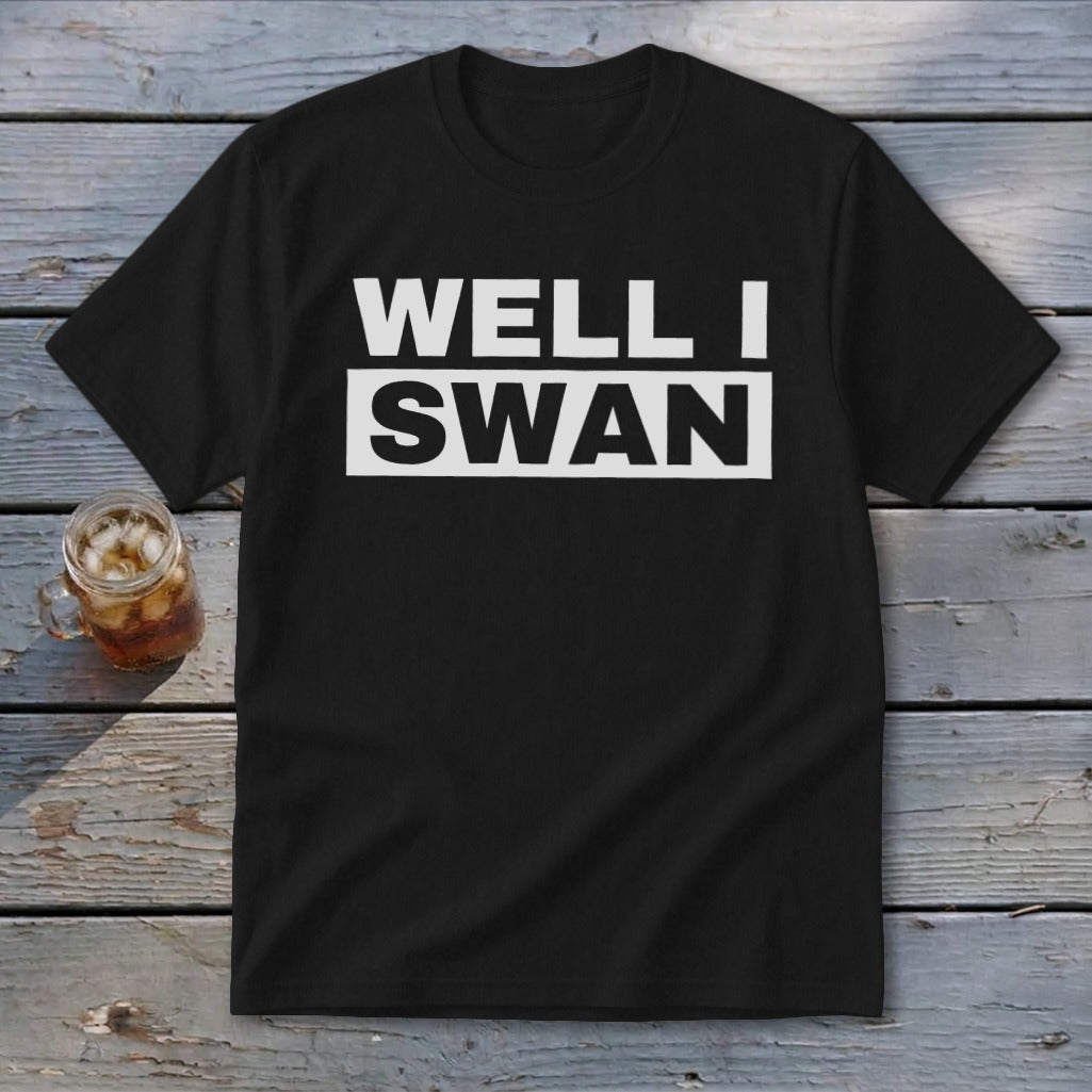 A black short-sleeve t-shirt with white text reading WELL I SWAN lies on a weathered wooden surface next to a glass of iced tea.