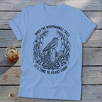 Light blue crewneck t-shirt featuring a black graphic of a bird on a wooden post, surrounded by corn stalks and ferns, with the text WHEN THE WHIPPOORWILL CALLS IT'S TIME TO PLANT CORN.