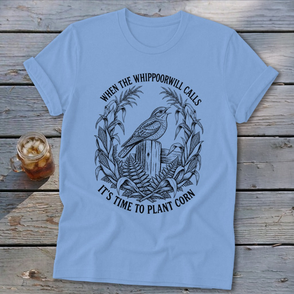 Light blue crewneck t-shirt featuring a black graphic of a bird on a wooden post, surrounded by corn stalks and ferns, with the text WHEN THE WHIPPOORWILL CALLS IT'S TIME TO PLANT CORN.