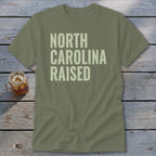 Olive green t-shirt with "NORTH CAROLINA RAISED" in light green text.