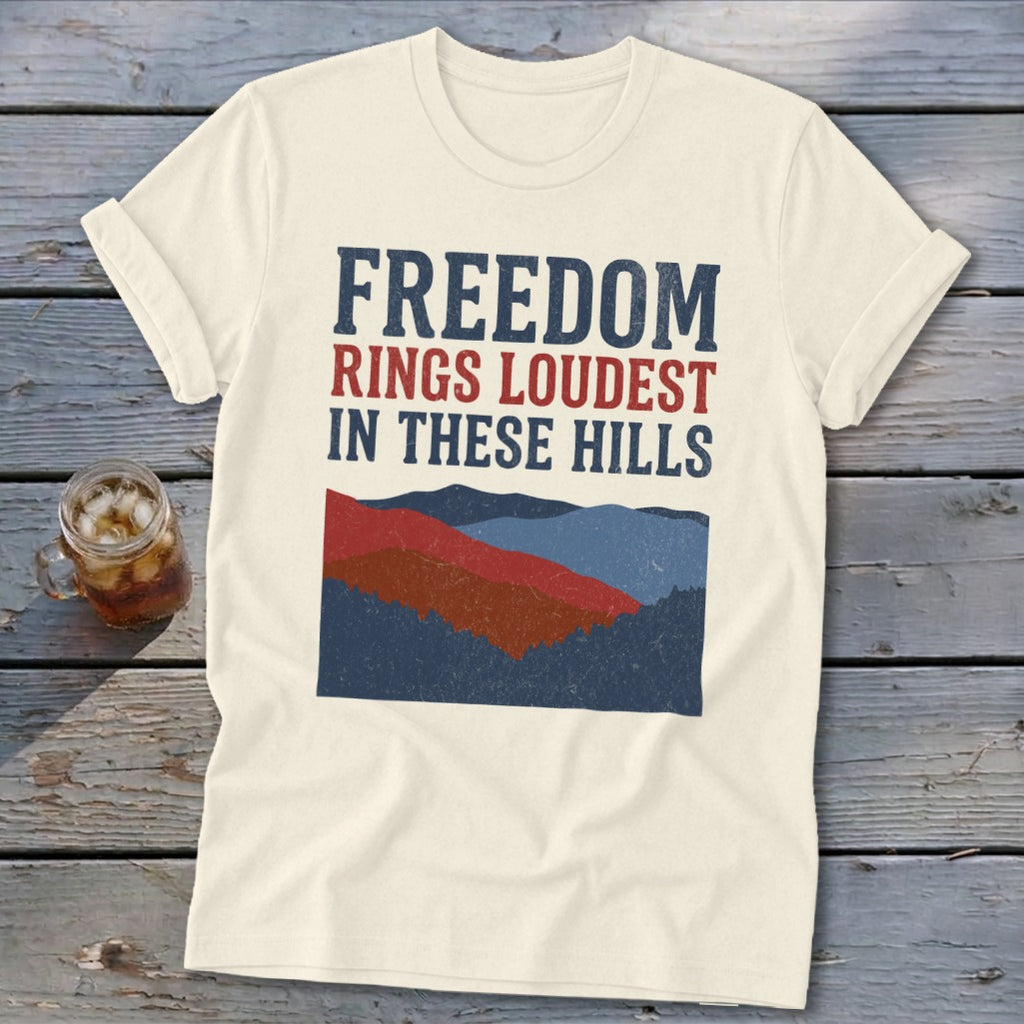 Cream t-shirt featuring distressed blue and red text that reads Freedom Rings Loudest in These Hills above a mountain graphic.
