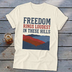 Cream t-shirt featuring distressed blue and red text that reads Freedom Rings Loudest in These Hills above a mountain graphic.