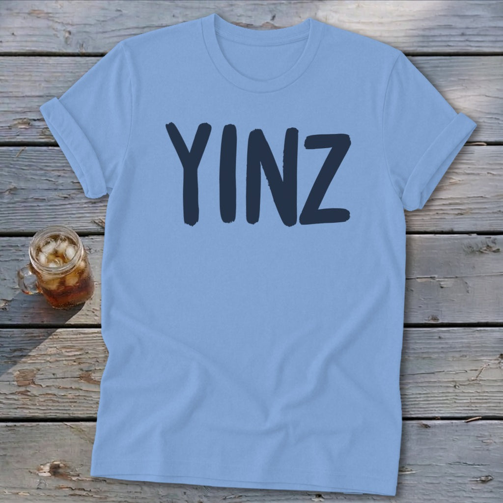 Light blue t-shirt with "YINZ" text in dark blue, laid on wooden planks.