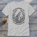 A light gray t-shirt featuring a black graphic of a bird perched on a wooden post, surrounded by corn stalks and the text "When the whippoorwill calls it's time to plant corn."
