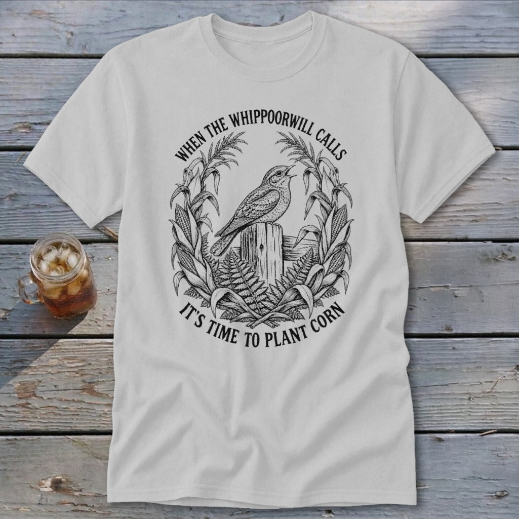 A light gray t-shirt featuring a black graphic of a bird perched on a wooden post, surrounded by corn stalks and the text "When the whippoorwill calls it's time to plant corn."