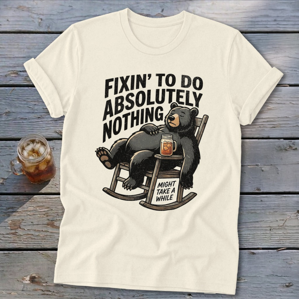 A cream-colored t-shirt with a graphic of a bear lounging in a rocking chair holding a drink, accompanied by the text Fixin to Do Absolutely Nothing and Might Take a While.