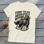 A cream-colored t-shirt with a graphic of a bear lounging in a rocking chair holding a drink, accompanied by the text Fixin to Do Absolutely Nothing and Might Take a While.