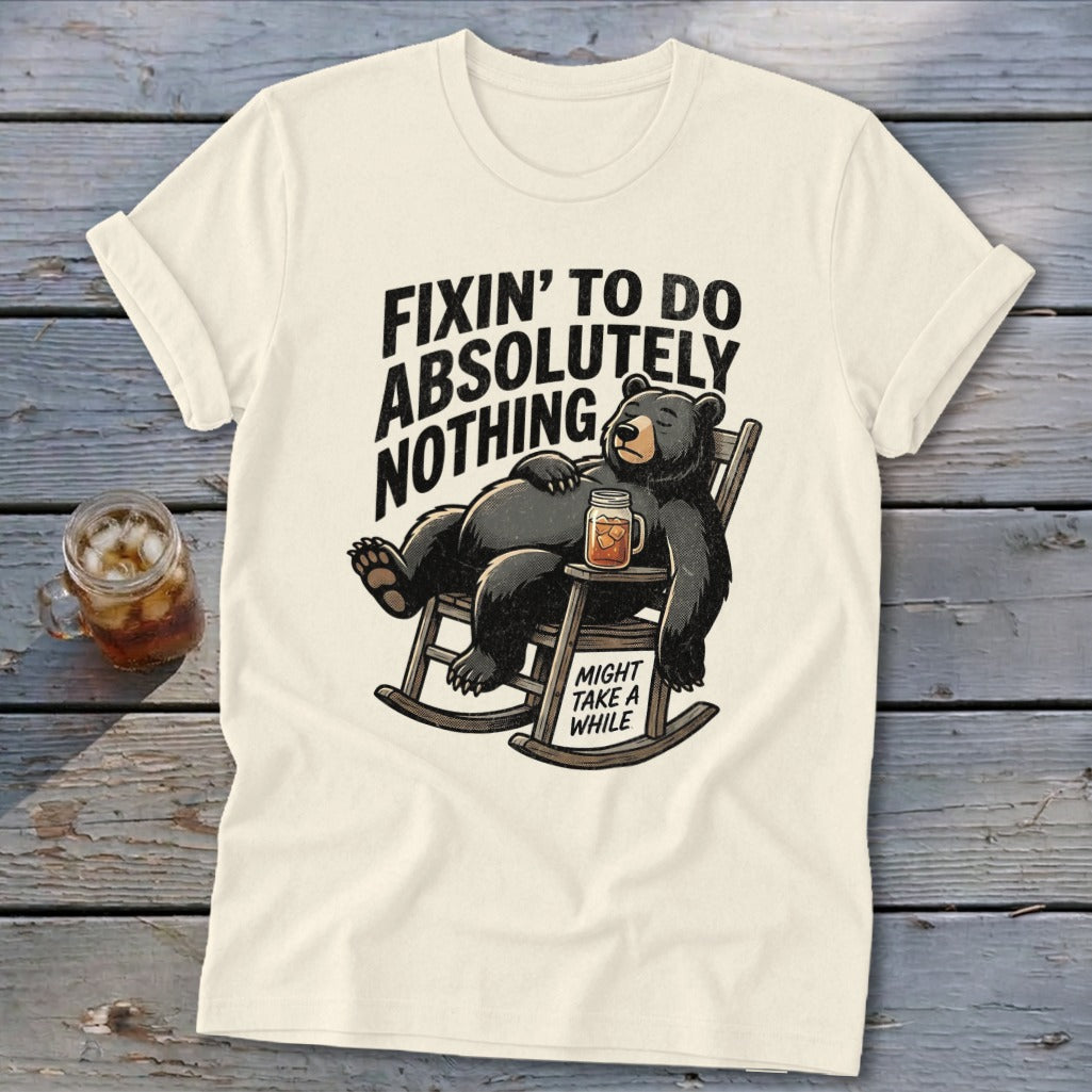 A cream-colored t-shirt with a graphic of a bear lounging in a rocking chair holding a drink, accompanied by the text Fixin to Do Absolutely Nothing and Might Take a While.