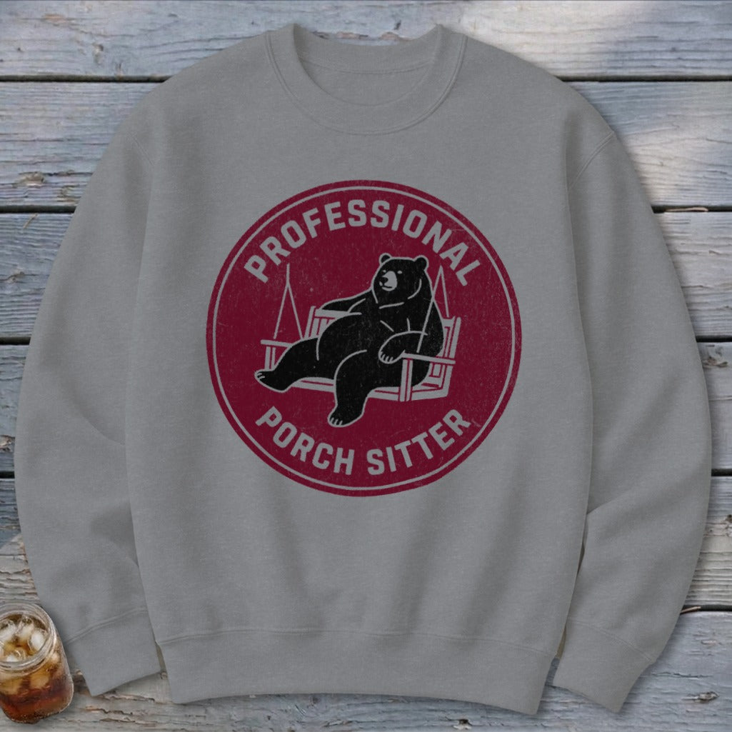 A gray crewneck sweatshirt features a circular maroon graphic of a bear relaxing on a porch swing, with the text "PROFESSIONAL PORCH SITTER."