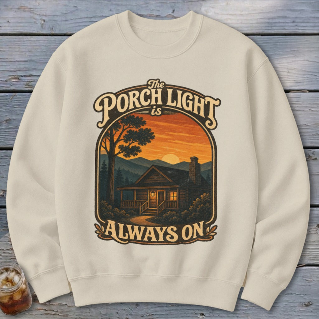 Cream sweatshirt with a graphic of a log cabin at sunset with text "The Porch Light is Always On."