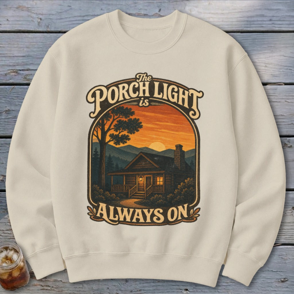 Cream sweatshirt with a graphic of a log cabin at sunset with text "The Porch Light is Always On."