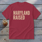 Crimson t-shirt with "MARYLAND RAISED" text in tan, next to a glass of iced beverage.