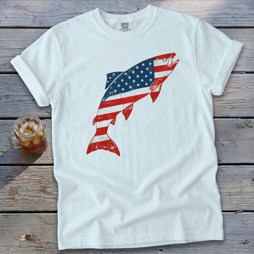 A white t-shirt featuring a jumping fish graphic patterned with the American flag on a wooden background.
