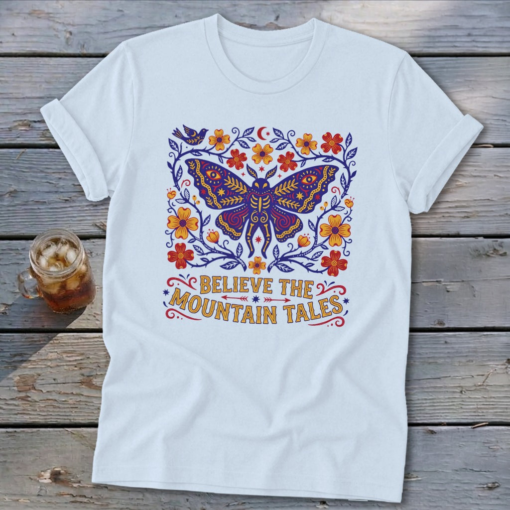 White t-shirt with a stylized butterfly, flowers, and text "Believe the Mountain Tales".