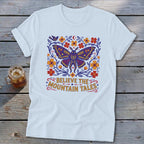 White t-shirt with a stylized butterfly, flowers, and text "Believe the Mountain Tales".