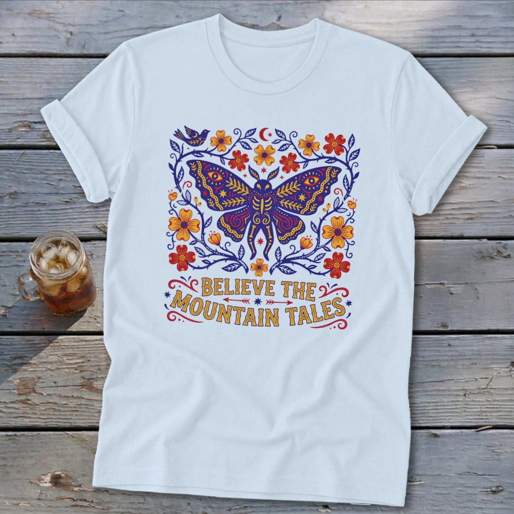White t-shirt with a stylized butterfly, flowers, and text "Believe the Mountain Tales".