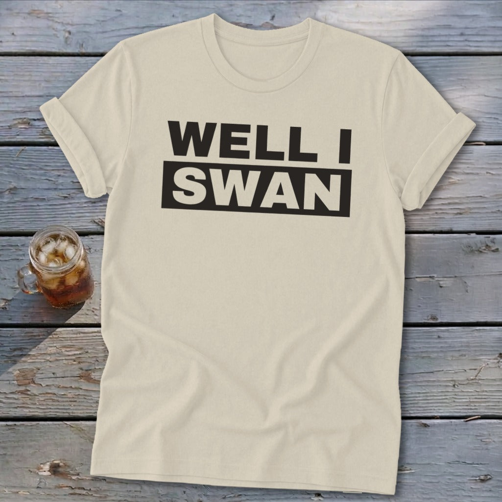 A cream-colored t-shirt with rolled sleeves lies on a wooden surface, featuring black text that reads WELL I SWAN.