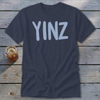 Navy t-shirt with light blue "YINZ" text, glass of iced drink beside it.