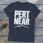 A dark blue t-shirt with rolled sleeves lies flat on a wooden surface, featuring the white text PERT NEAR above a simple mountain outline.