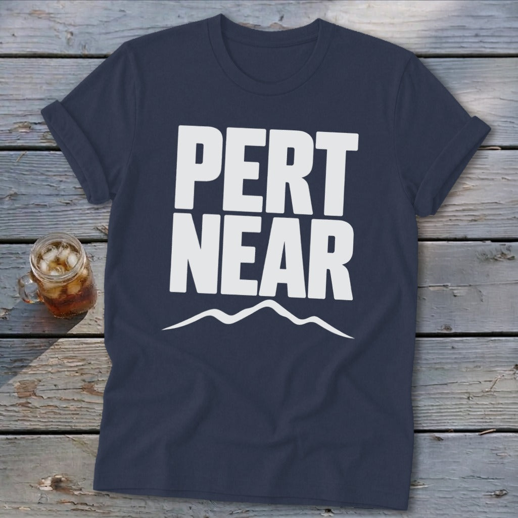 A dark blue t-shirt with rolled sleeves lies flat on a wooden surface, featuring the white text PERT NEAR above a simple mountain outline.