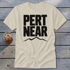 A cream-colored t-shirt rests on a wooden surface, featuring bold black text reading PERT NEAR above a mountain silhouette.