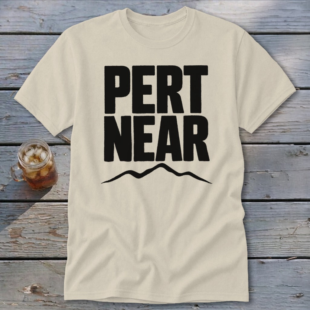 A cream-colored t-shirt rests on a wooden surface, featuring bold black text reading PERT NEAR above a mountain silhouette.