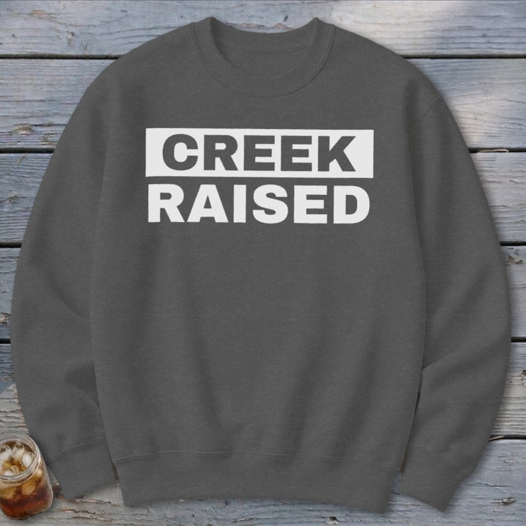 Dark grey crewneck sweatshirt with white text "CREEK RAISED" inside a white box.