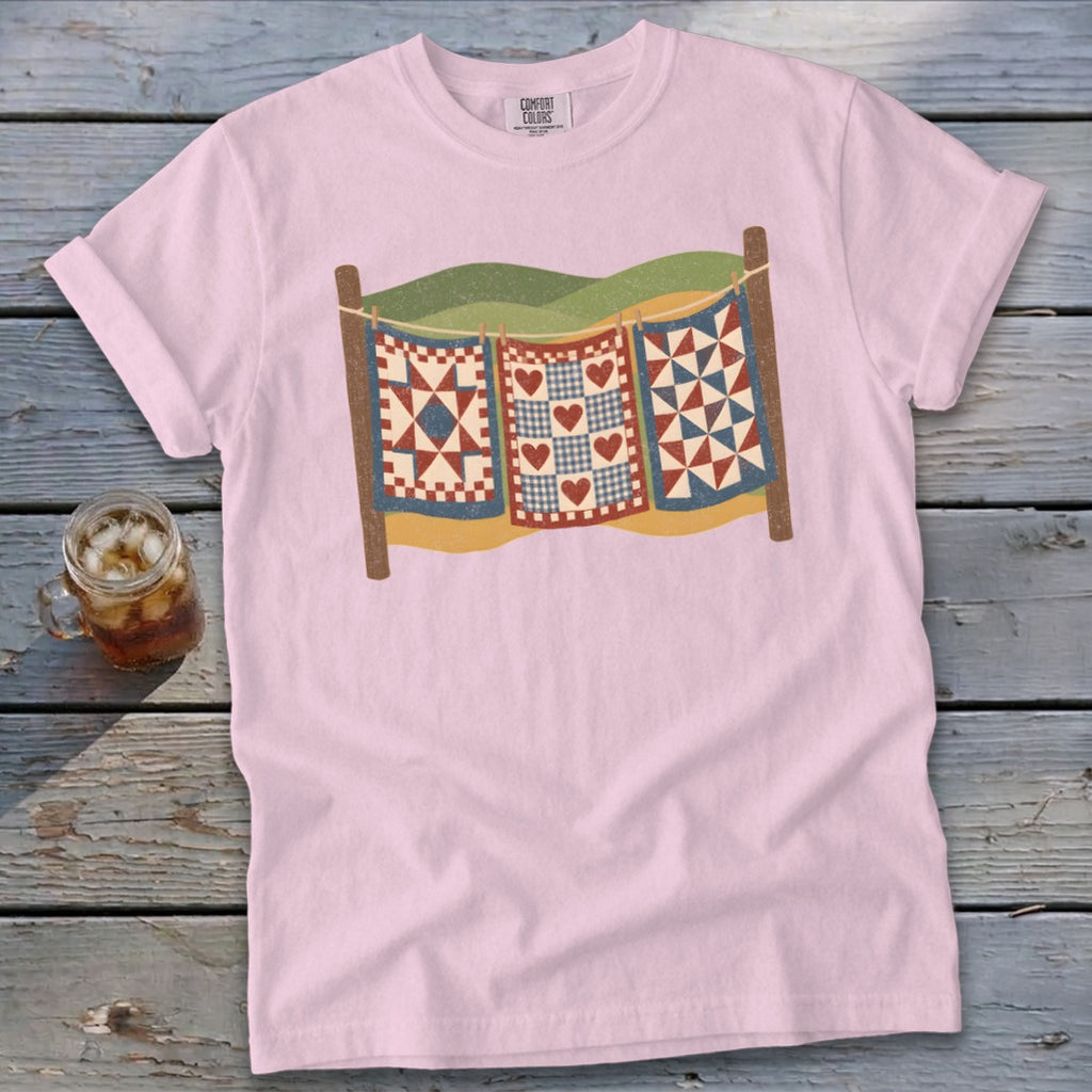 Light pink t-shirt featuring a graphic of three colorful quilts hanging on a clothesline against a wooden background.