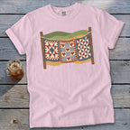 Light pink t-shirt featuring a graphic of three colorful quilts hanging on a clothesline against a wooden background.