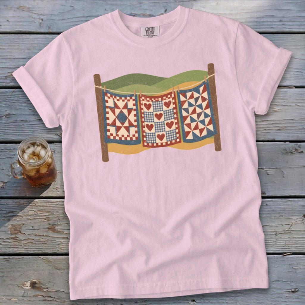 Light pink t-shirt featuring a graphic of three colorful quilts hanging on a clothesline against a wooden background.