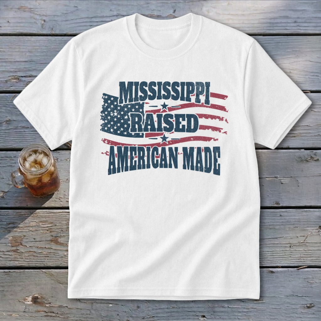 White t-shirt with distressed American flag graphic and text reading Mississippi Raised American Made on wooden background.