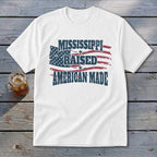White t-shirt with distressed American flag graphic and text reading Mississippi Raised American Made on wooden background.