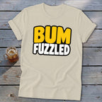 Beige short-sleeve t-shirt with rolled cuffs, featuring the black-outlined text BUM FUZZLED in yellow and white on a wooden background.