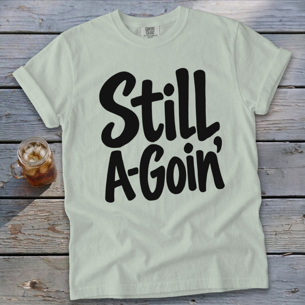 Light green t-shirt with black cursive text reading Still A-Goin' on a wooden background.